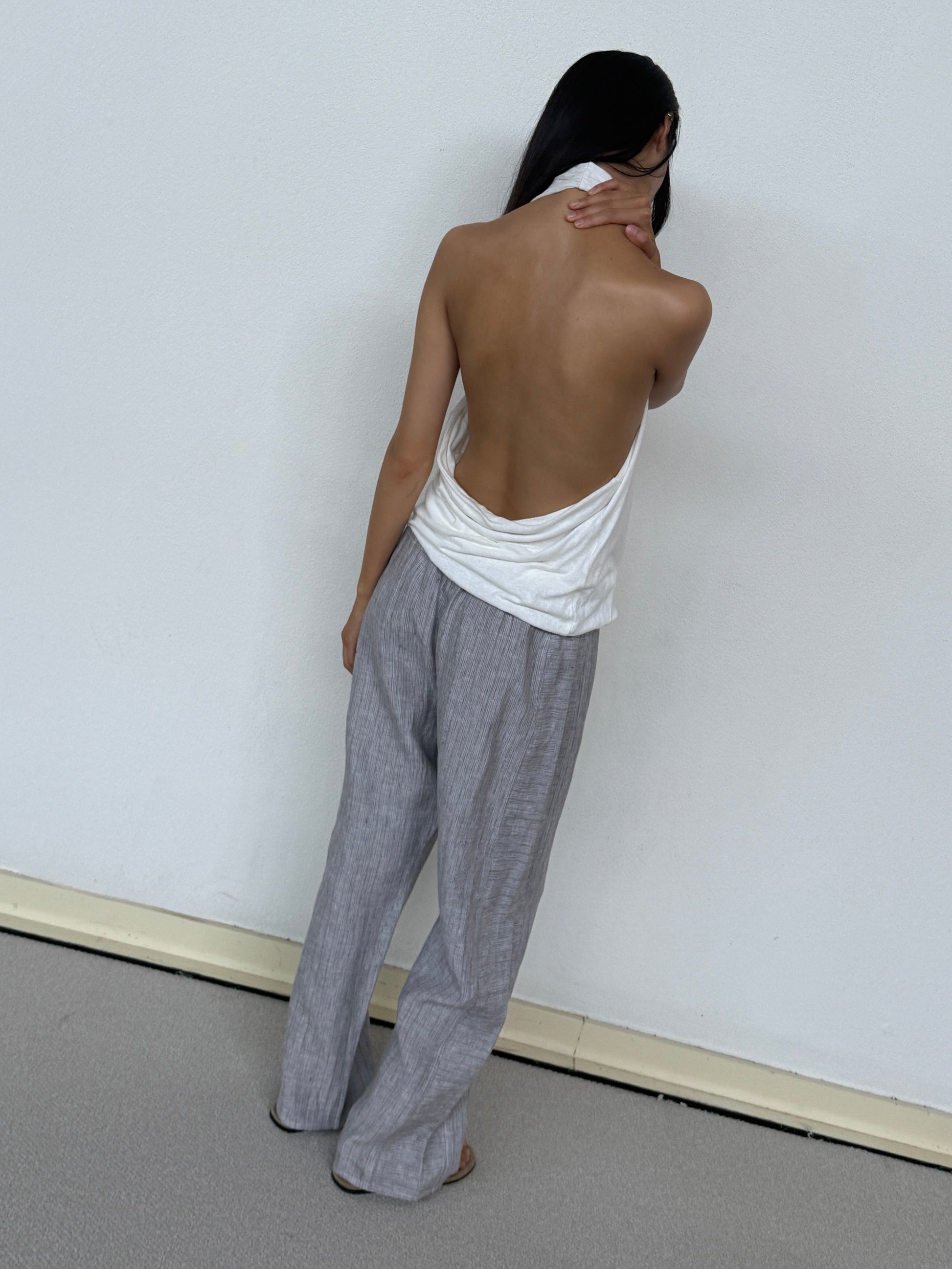 A person stands barefoot facing a white wall, wearing 0s Double Waist Linen Pant in Haze Blue and a backless white top, with one hand touching the back of their neck.