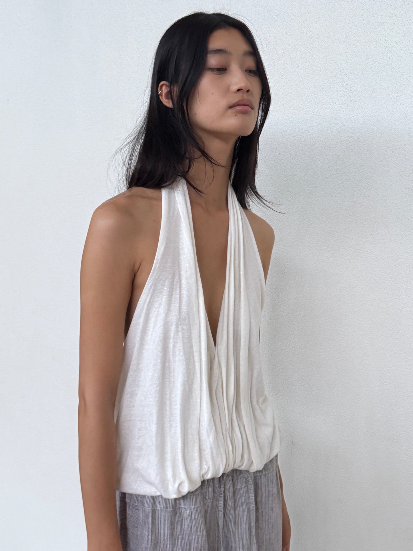 A person with long dark hair wears a sleeveless, draped white Jersey Top and gray pants, standing against a plain white wall and looking slightly down and to the side.