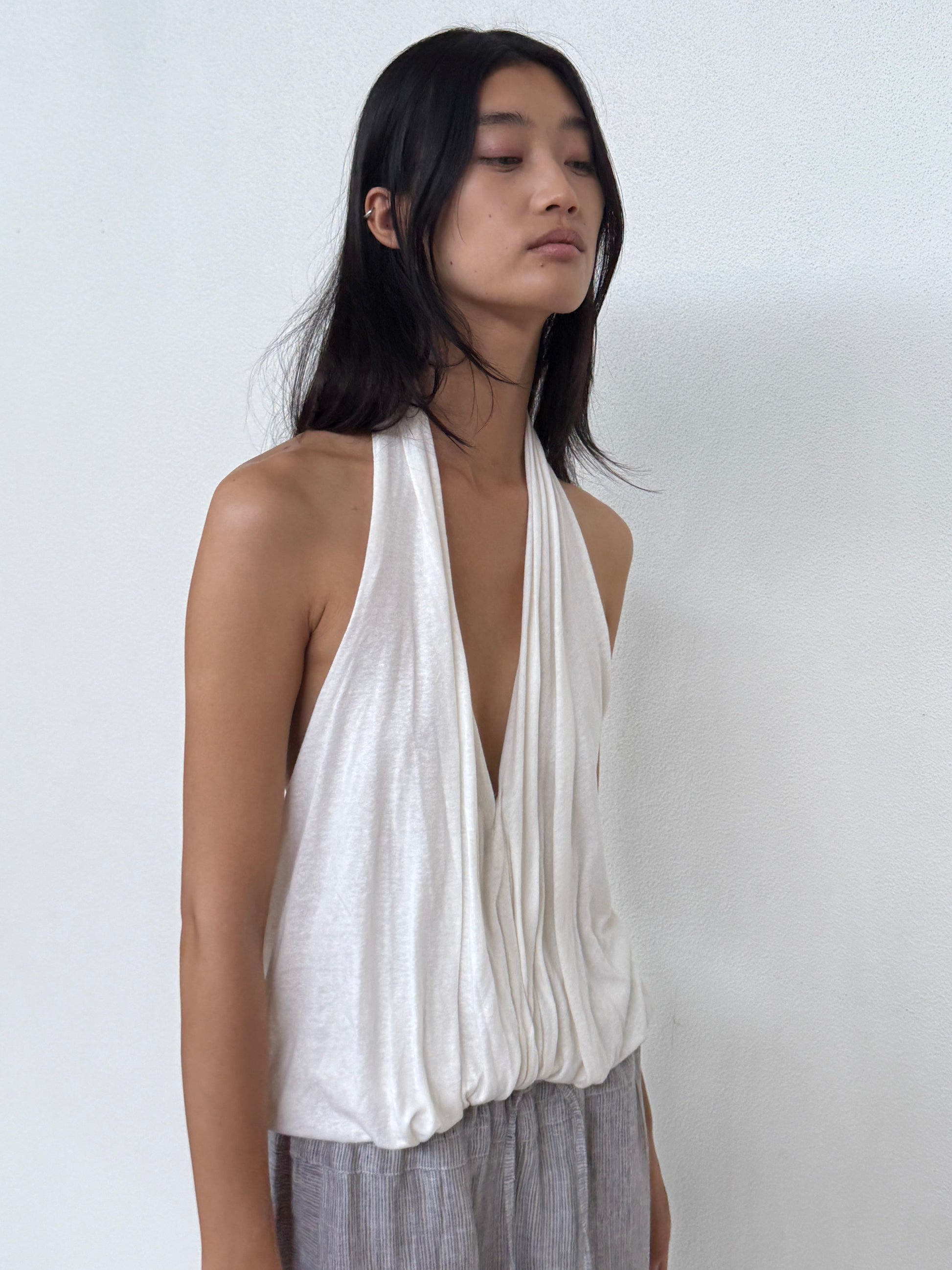 A person with long dark hair wears the 0 Jersey Top in white and light gray pants, standing against a plain white wall and gazing downward with a calm expression.