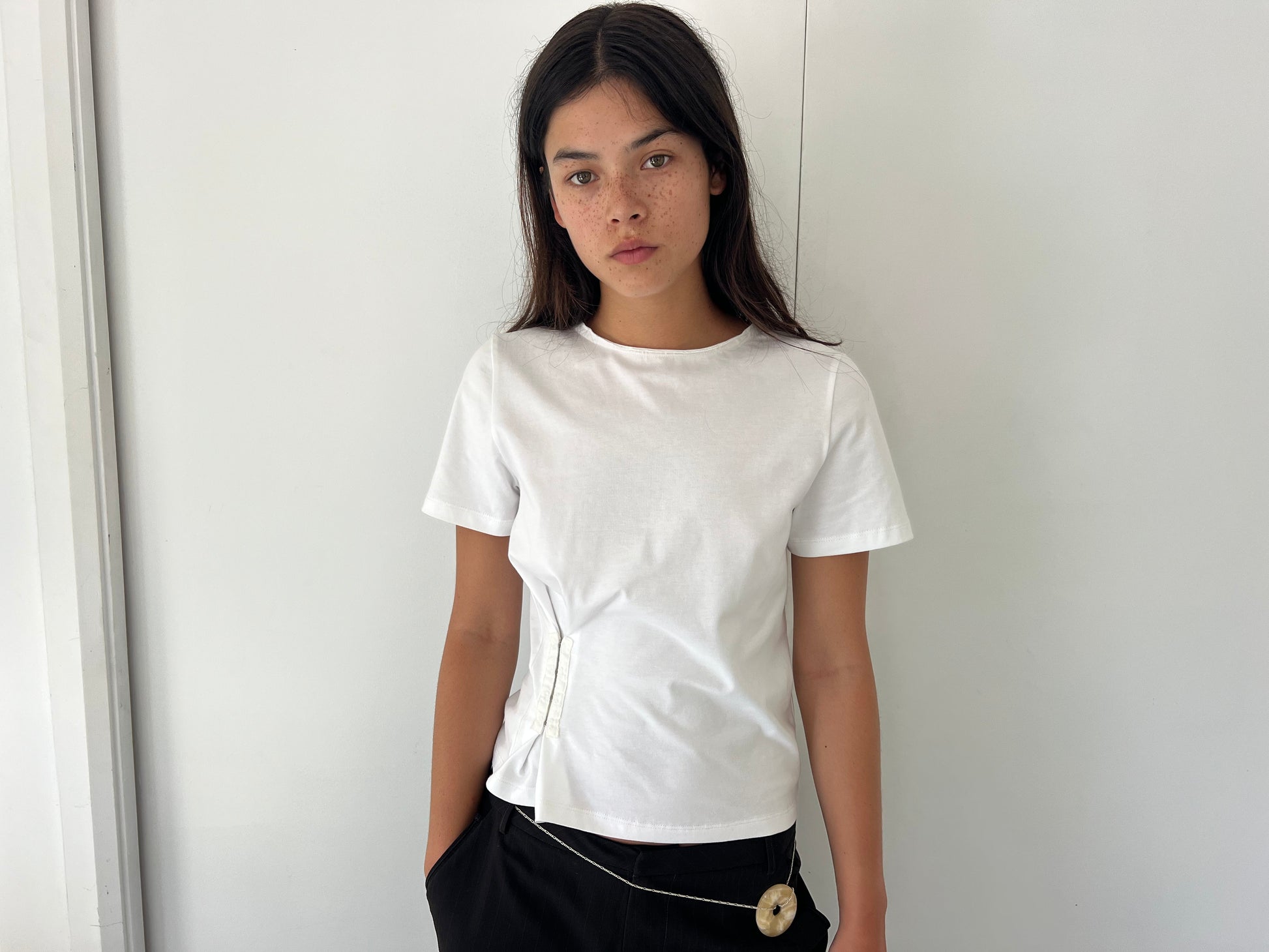 A young woman with long dark hair and freckles stands against a white wall, wearing the Pinch Tee - White by Deiji Studios and black pants, one hand in her pocket and a neutral expression.