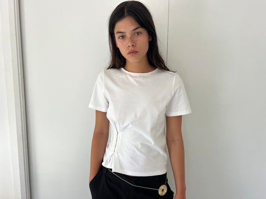A young woman with long dark hair and freckles stands against a plain white wall wearing the Pinch Tee - White from Deiji Studios and black pants, one hand in her pocket and a neutral expression.