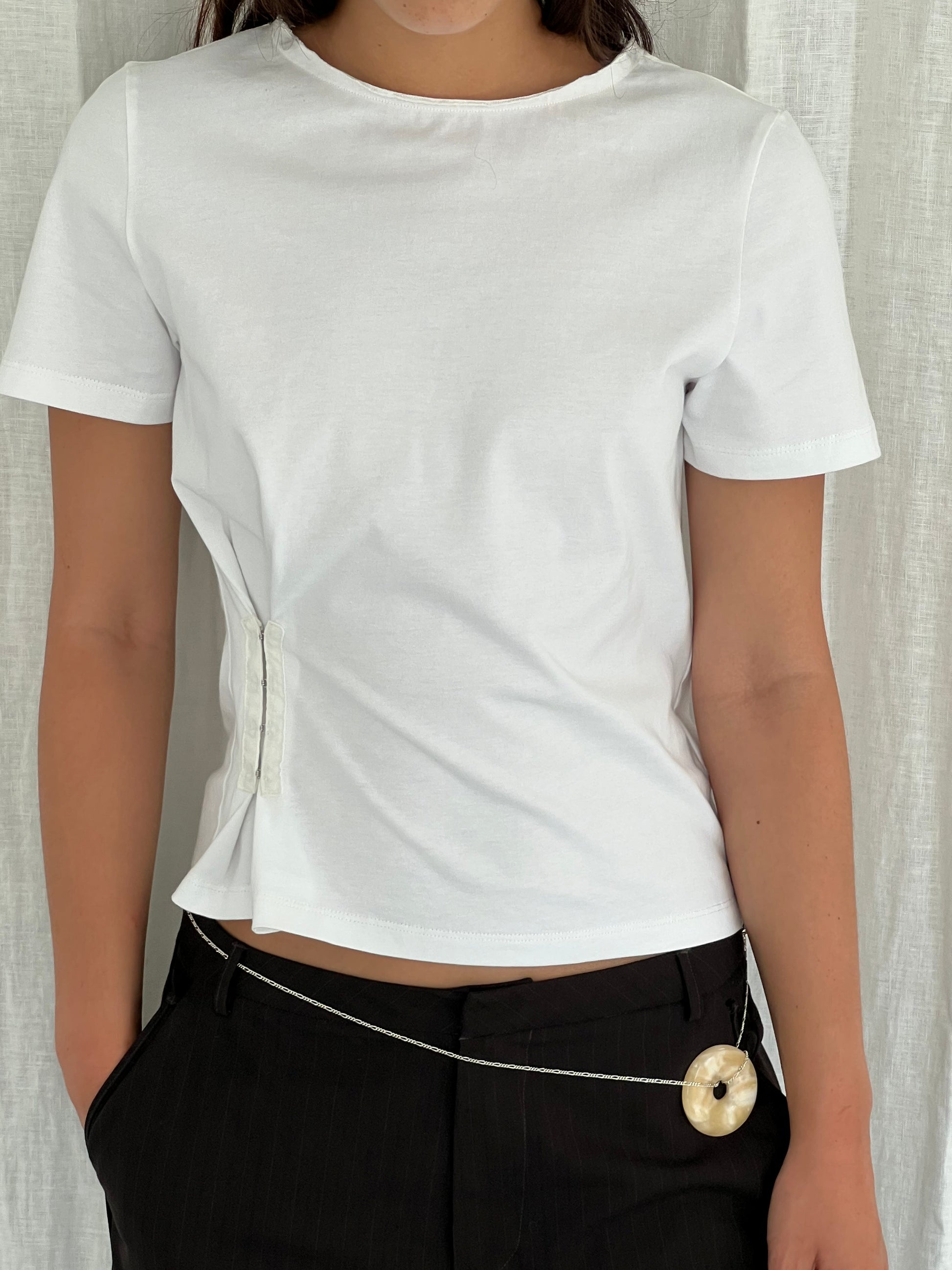 Someone wearing the Deiji Studios Pinch Tee - White, black pants, and a thin chain belt with a round light-colored pendant stands in front of a white curtain.