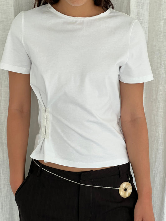 A person wears the Pinch Tee - White by Deiji Studios with black pants and a thin belt featuring a large round beige pendant. A white curtain serves as the background.