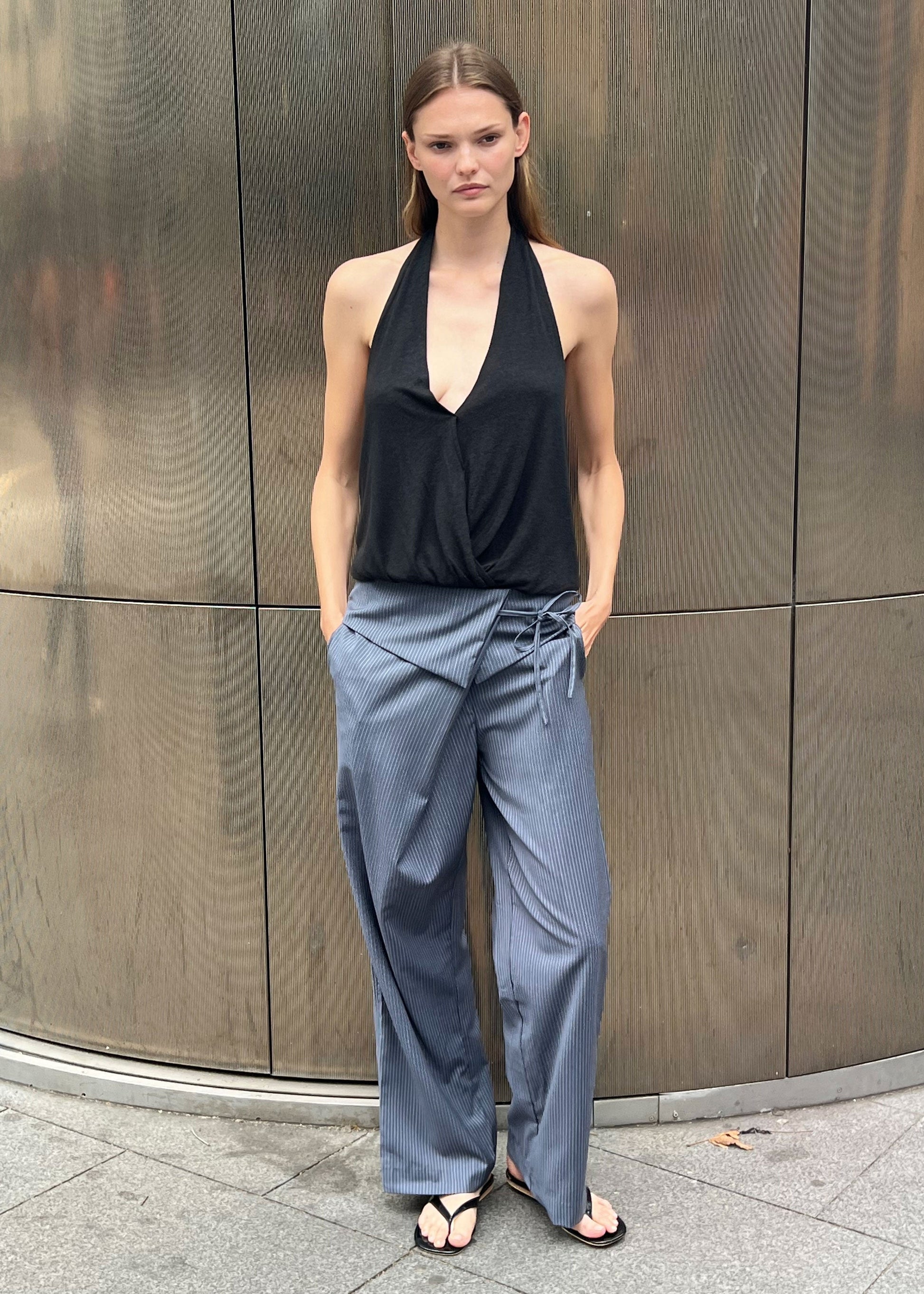 A woman stands outdoors by a metallic wall, wearing a black halter top and the Folded Wool Pant - Navy Stripe with her hands in her pockets and a neutral expression.