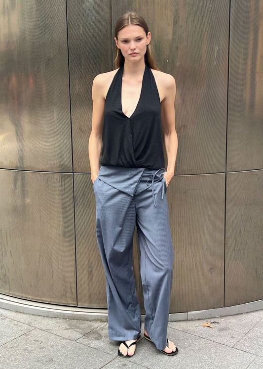 A woman stands outdoors against a metallic curved wall, wearing a black halter-neck top, black sandals, and the Folded Wool Pant—loose gray pinstripe trousers with a wrap detail. Her hands are in her pockets and she has a neutral expression.