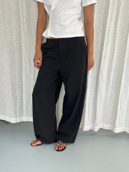 A person stands against a white curtain wearing a white short-sleeve top, Bind Pant - Charred Black Stripe by Deiji Studios, and black sandals. Their face is not visible in the image.