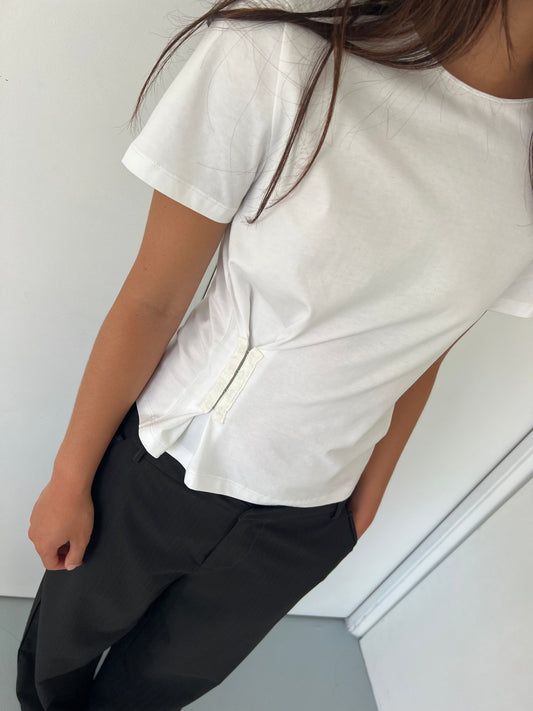 A person wearing the Pinch Tee - White by Deiji Studios, featuring side clasps, paired with black pants stands against a white wall with their head partially out of frame.