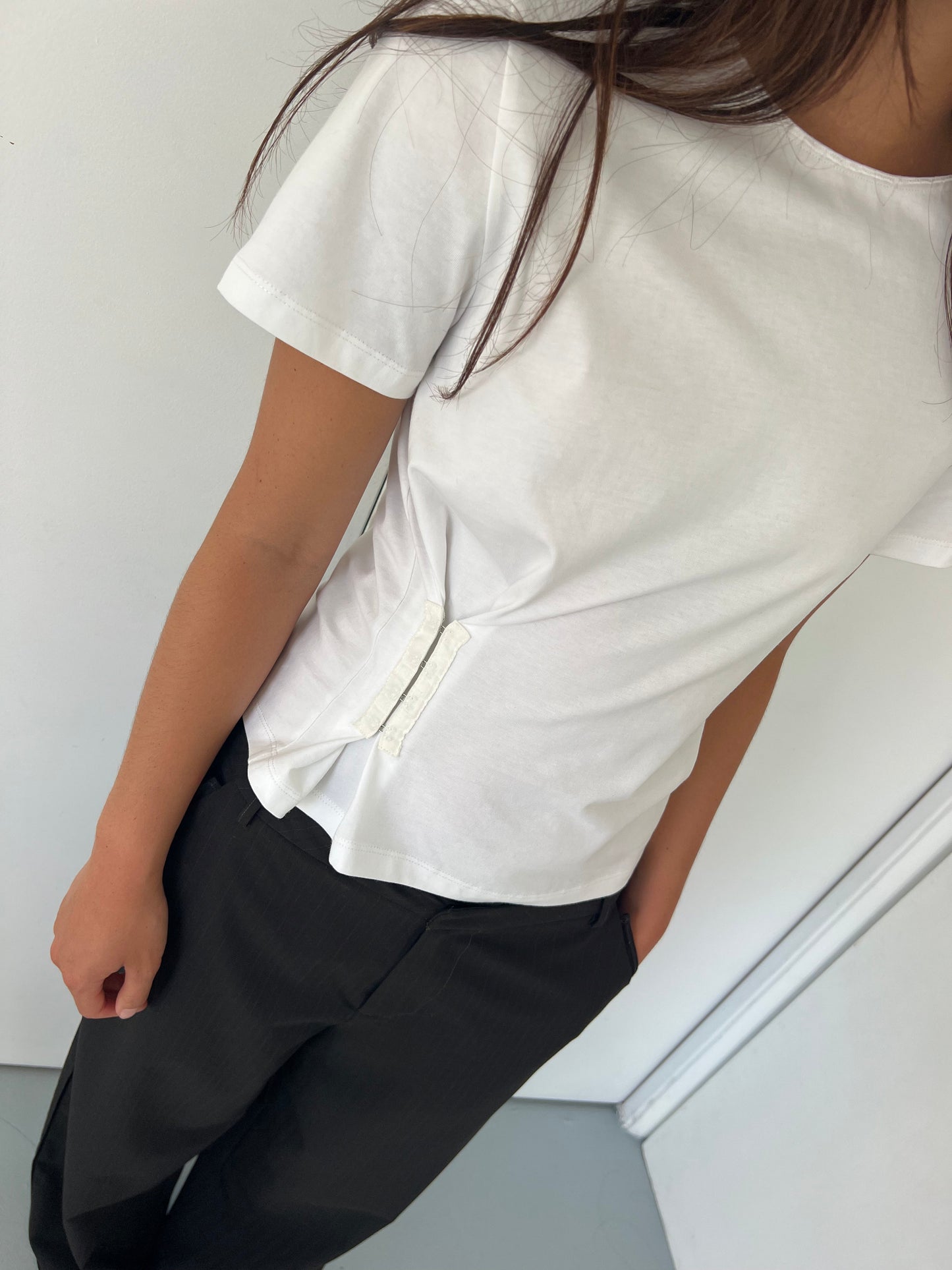 A person wears the Deiji Studios Pinch Tee - White, featuring a relaxed short sleeve design with two vertical side straps, paired with black pants near a white wall; their face is out of frame.