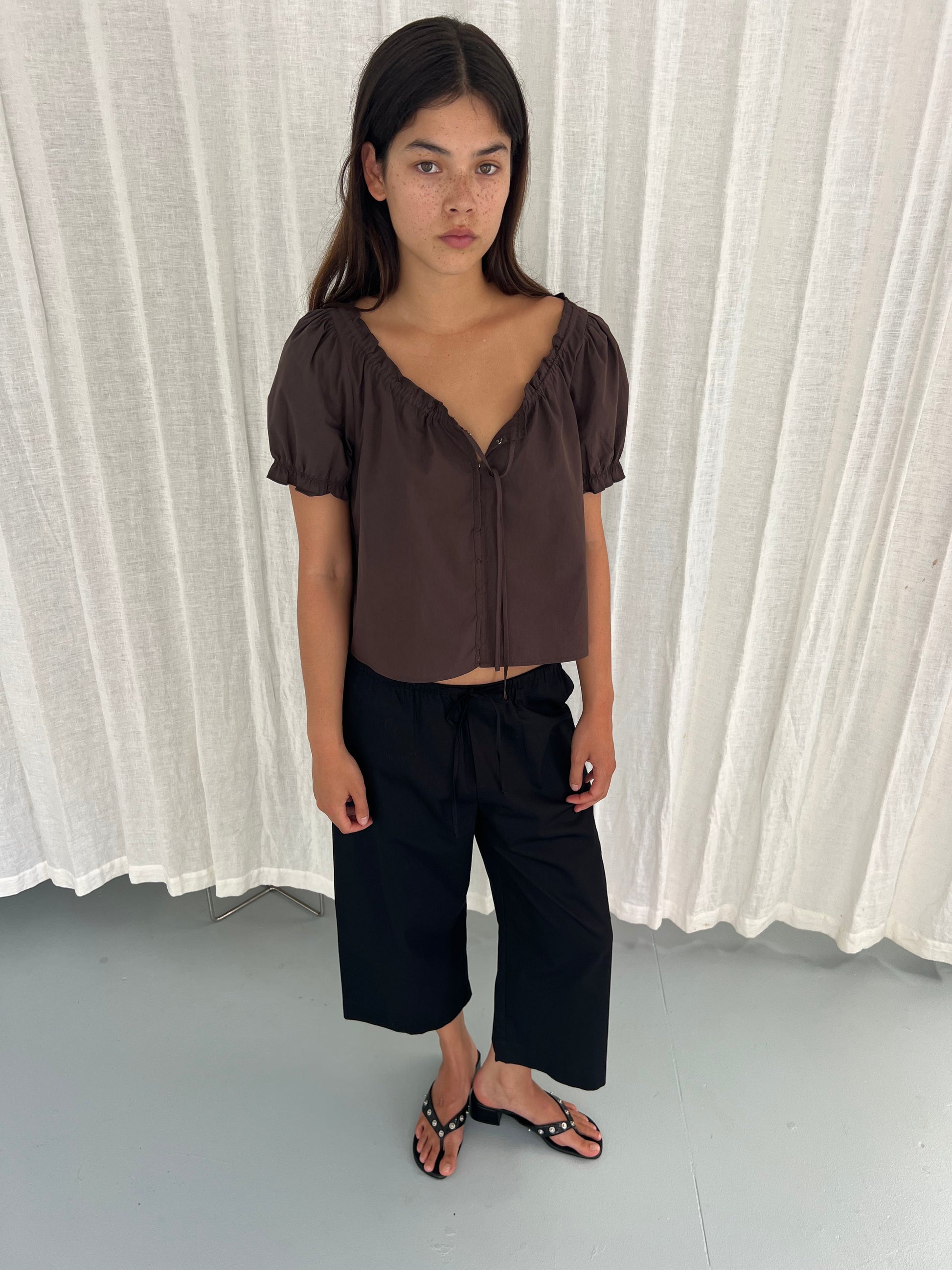 A young woman with long dark hair stands indoors on a light grey floor before white curtains, wearing a Deiji Studios Close Shirt in Sepia, black wide-leg pants, and black sandals. She gazes slightly to the side with a neutral expression.