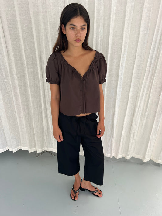 A young woman with long dark hair stands before white curtains, wearing the Close Shirt - Sepia by Deiji Studios with a cropped, relaxed fit, styled with black wide-leg pants and sandals. She gazes to the side with a neutral expression.