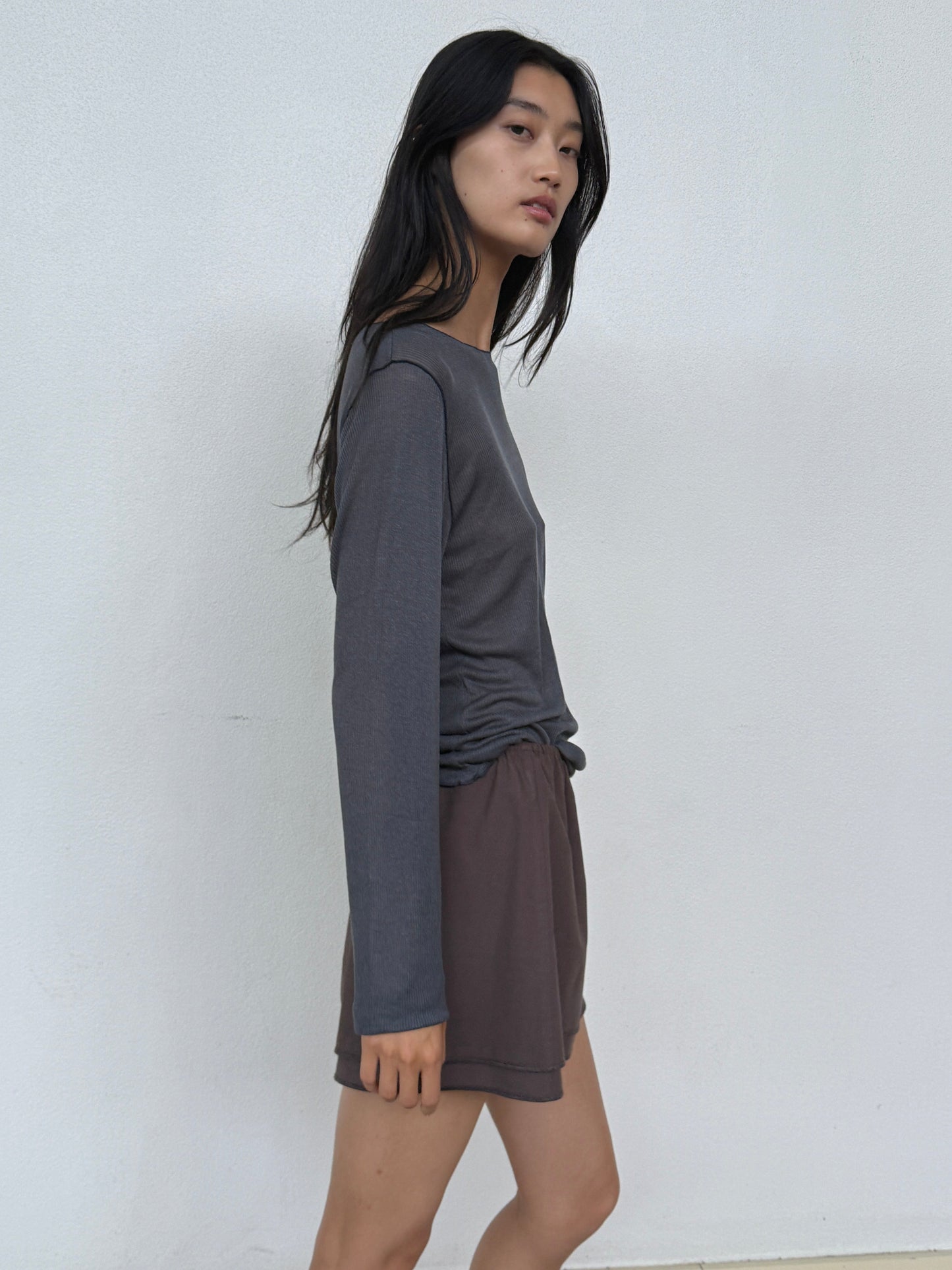 A person with long dark hair stands against a white wall, wearing a relaxed 0 Knit Long Sleeve - Steel and brown shorts, looking sideways at the camera.