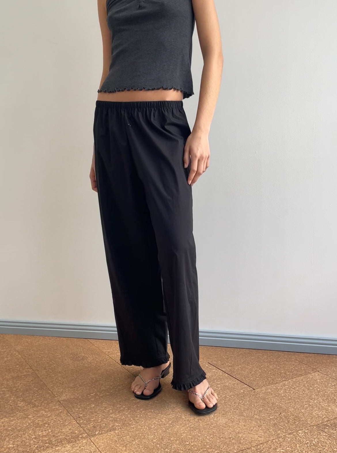 A person stands indoors on a cork floor, facing a white wall and wearing the Flounce Pant - Black from Deiji Studios, styled with a sleeveless dark crop top for an effortlessly chic look.