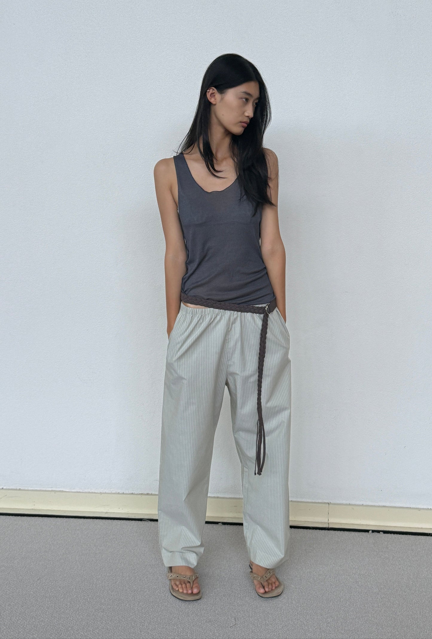 A woman with long black hair stands by a white wall, wearing the Knit Tank - Steel from 0, loose light gray pants with a thin belt, and sandals. She looks to the side with her hands in her pockets.