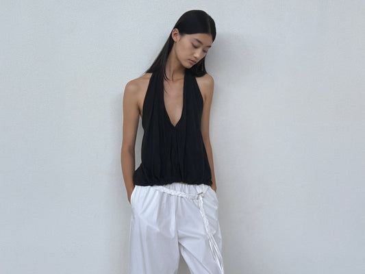 A person with long dark hair wears the Jersey Top - Black by 0, a sleeveless V-neck piece, paired with loose white pants and a braided belt. They stand by a plain light wall, hands in pockets, gazing slightly downward.