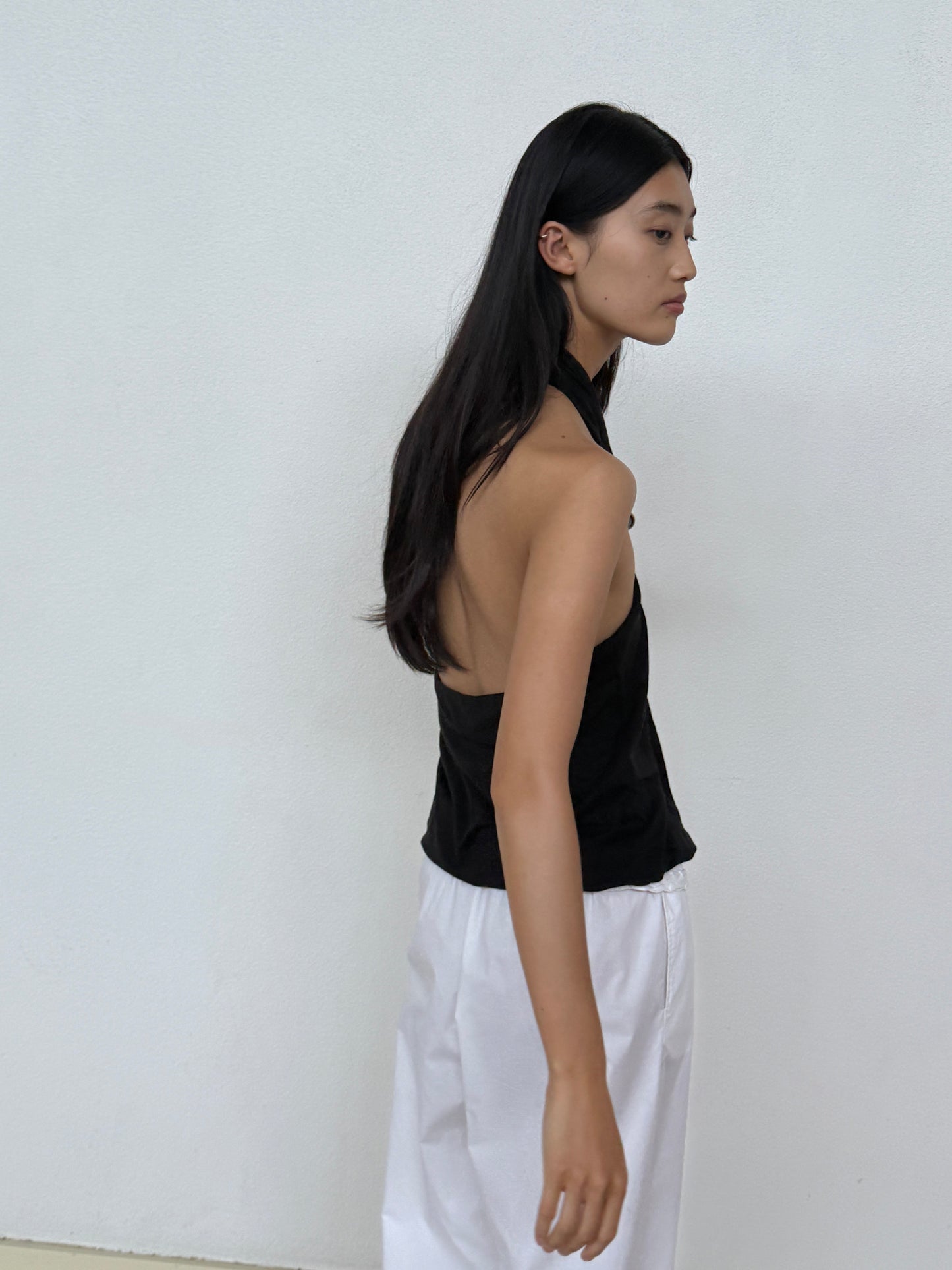 A woman with long dark hair stands against a white wall, wearing the Jersey Top - Black by 0 and loose white pants with a relaxed fit, looking to her left with a neutral expression.