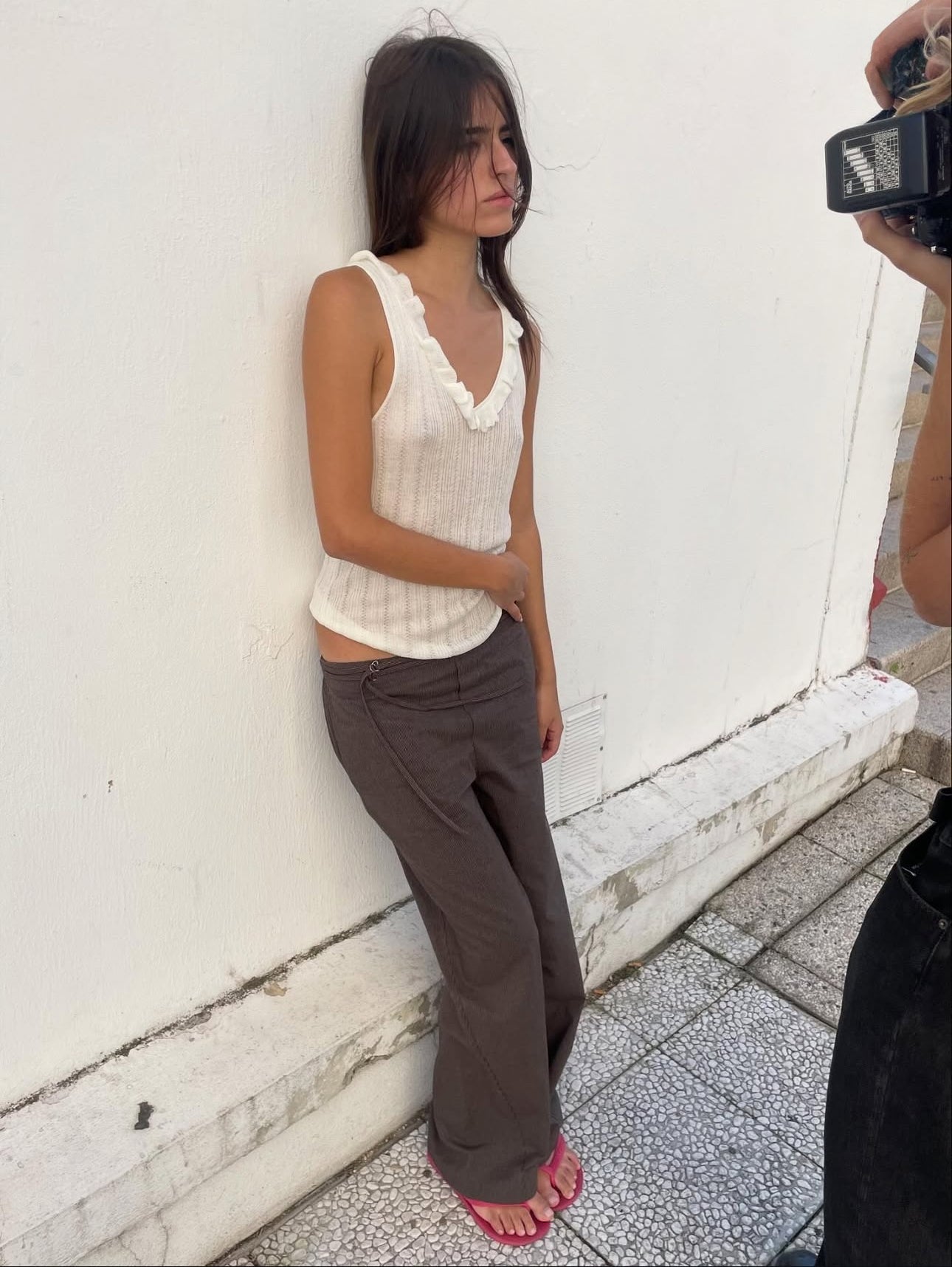 A woman with long dark hair, dressed in a white sleeveless top and Double Belted Pant - Brown Stripe, stands against a white wall as someone photographs her. Pink sandals finish her effortlessly chic look.