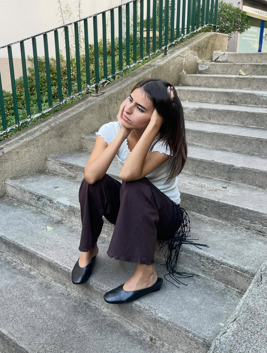 A young woman with long dark hair sits on outdoor concrete steps, resting her elbows on her knees and her head in her hands. She wears a Deiji Studios white shirt and the Flounce Pant - Umber with black flats.
