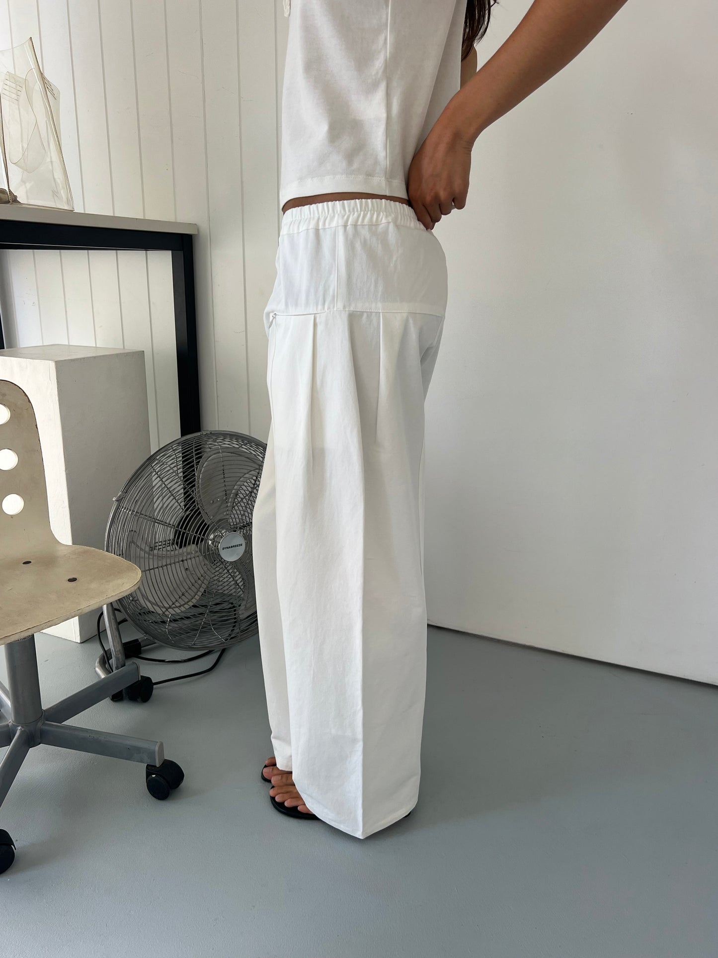 Someone stands indoors in a minimalist room, adjusting the fit of the Pleat Pant - White with an elastic waistband and loose pleats. Only part of their upper body is visible, along with a fan, chair, and desk in the background.