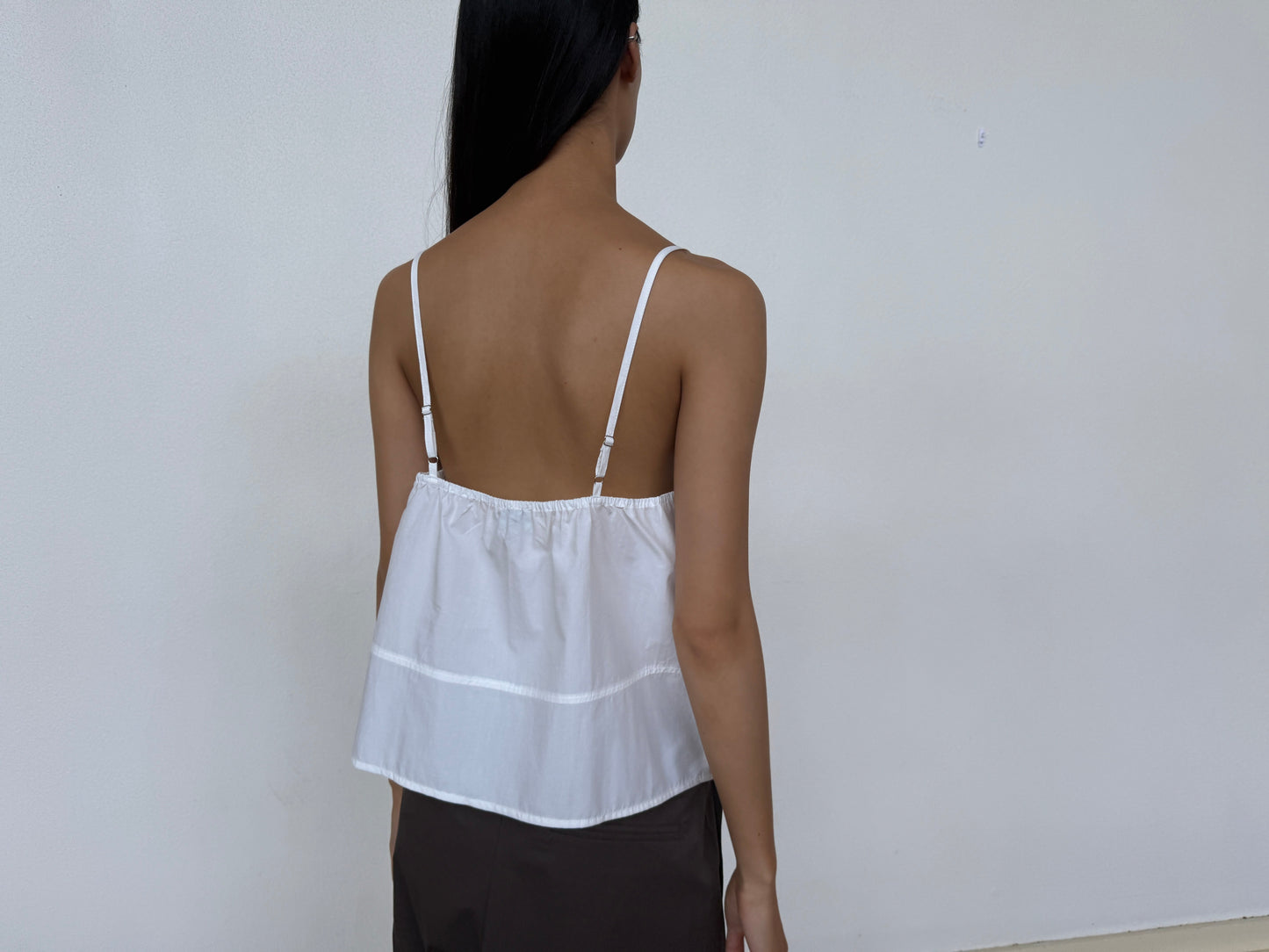 A person with long dark hair, seen from behind, wears a Seam Top - White by 0, made of organic cotton, paired with dark bottoms and standing before a plain light wall.