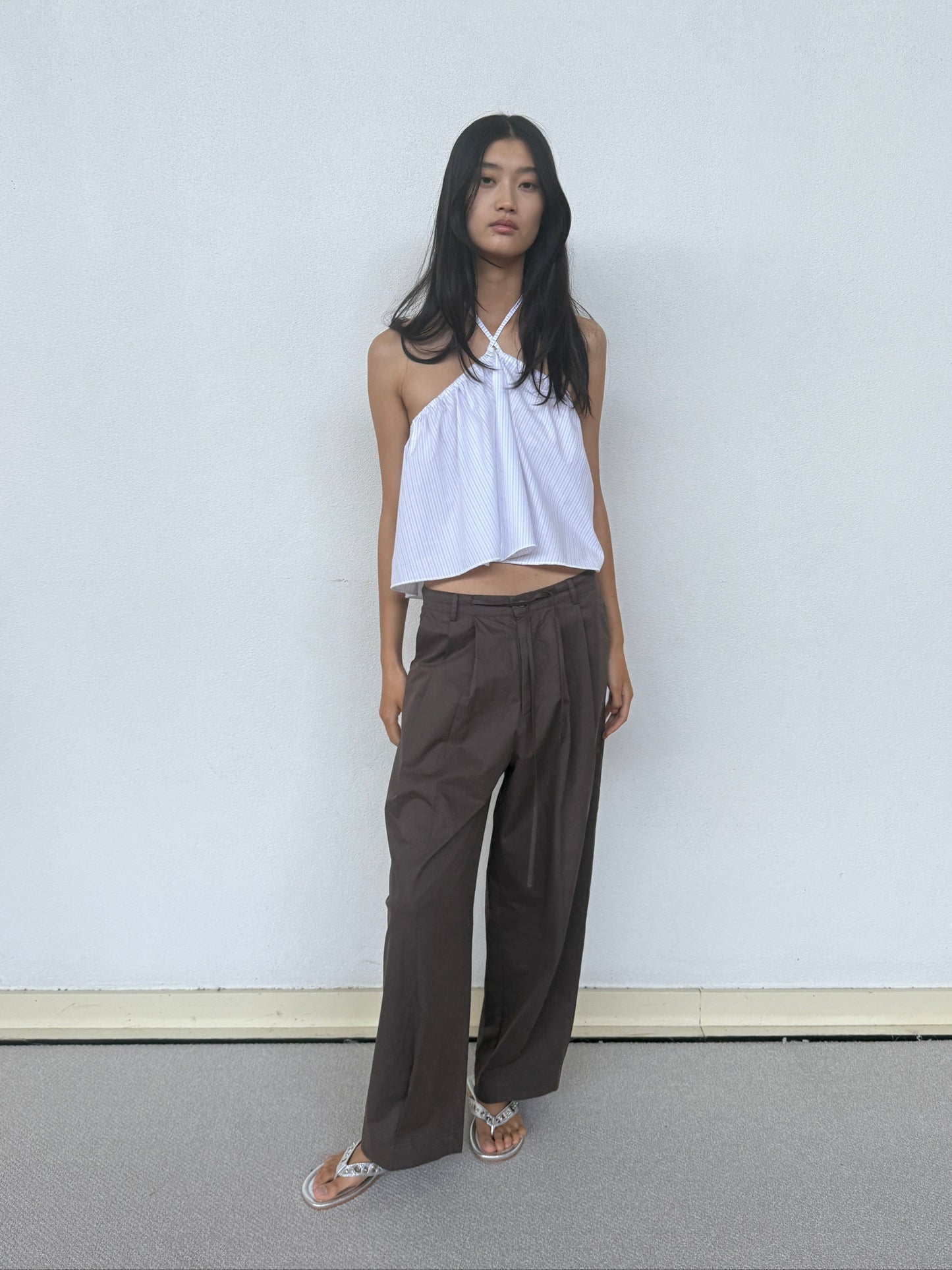 A woman with long black hair stands against a light wall, wearing a white halter neck sleeveless top, Turn Pant - Cocoa by Deiji Studios with an adjustable drawcord, and sandals. Her arms are at her sides and she looks relaxed.