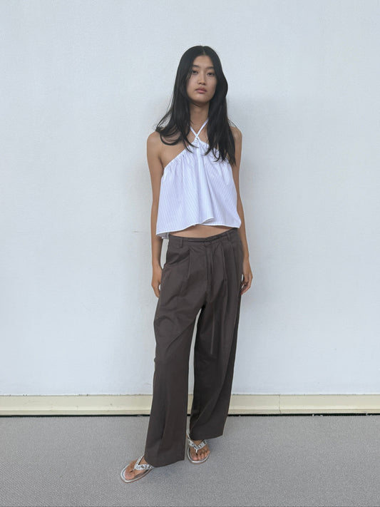 A woman with long black hair stands against a light wall, wearing a white halter neck sleeveless top, Turn Pant - Cocoa by Deiji Studios with an adjustable drawcord, and sandals. Her arms are at her sides and she looks relaxed.