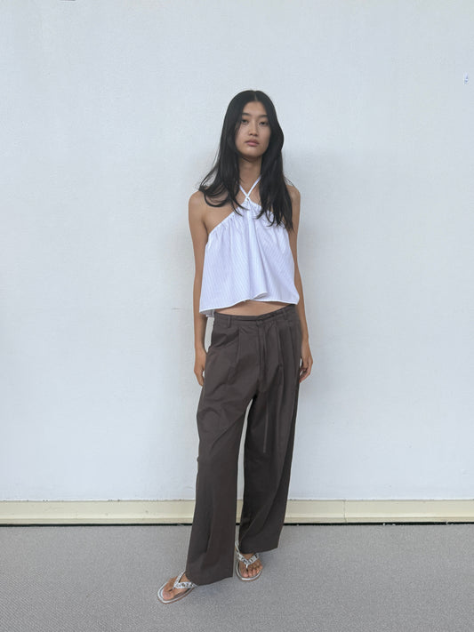 A young woman with long dark hair and a relaxed expression stands against a plain light wall, wearing a sleeveless white halter top, 0 Turn Pant in Cocoa, and sandals.