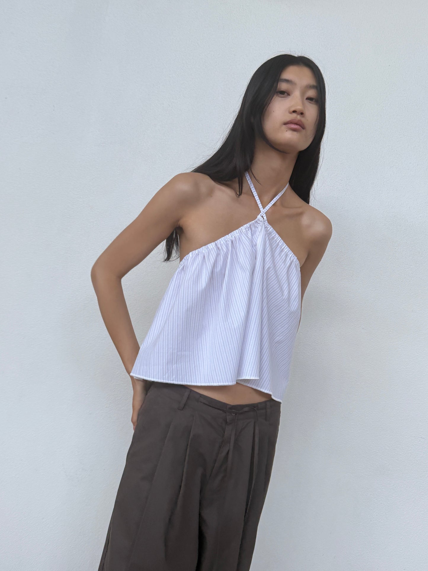 A woman with long, dark hair stands confidently against a white wall, wearing the 0 Napkin Top - Pacific Stripe paired with high-waisted, wide-leg brown pants.