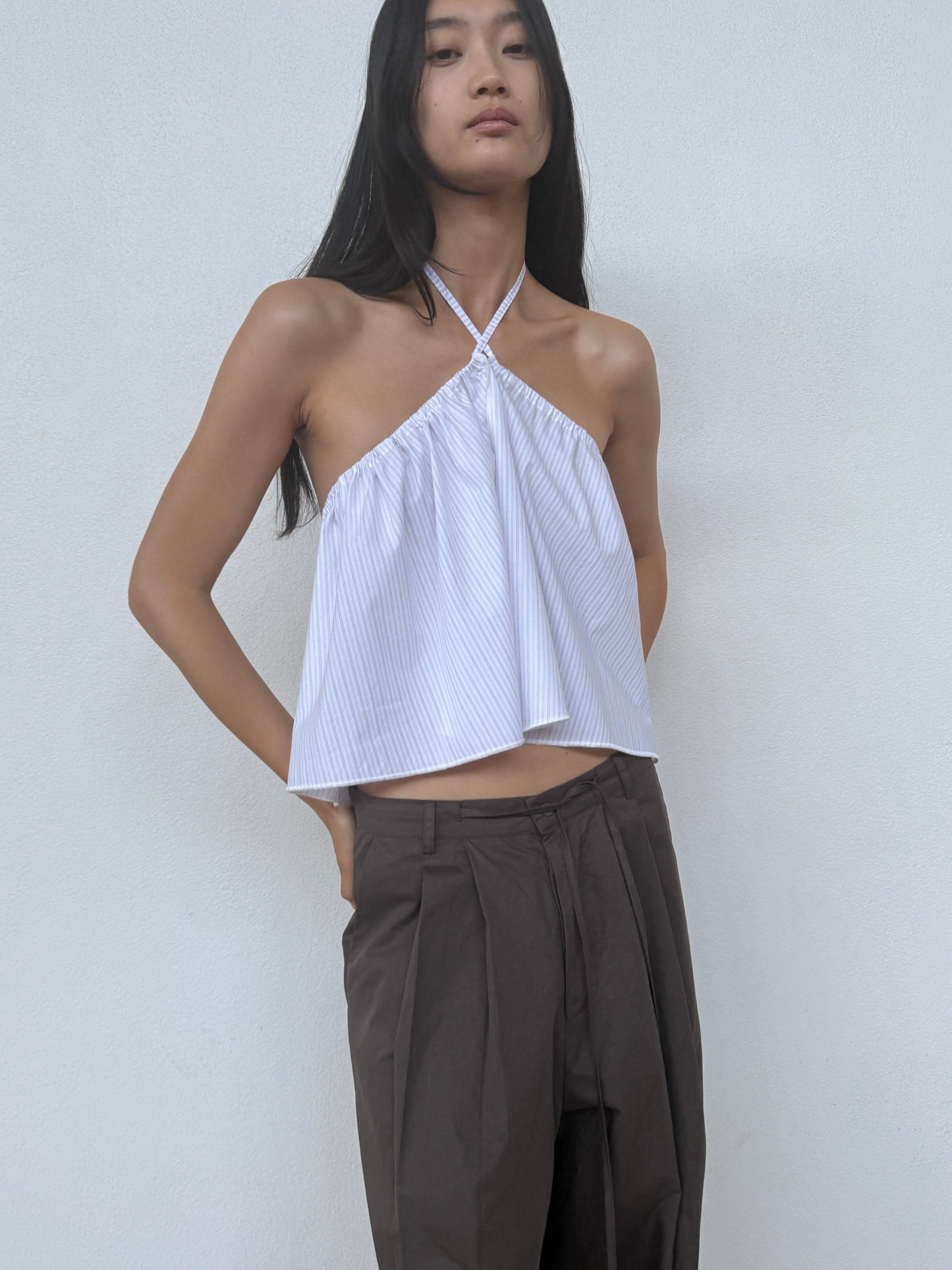 A person with long dark hair wears the Turn Pant - Cocoa by 0 with a white halter-neck top, standing against a plain light-colored wall.