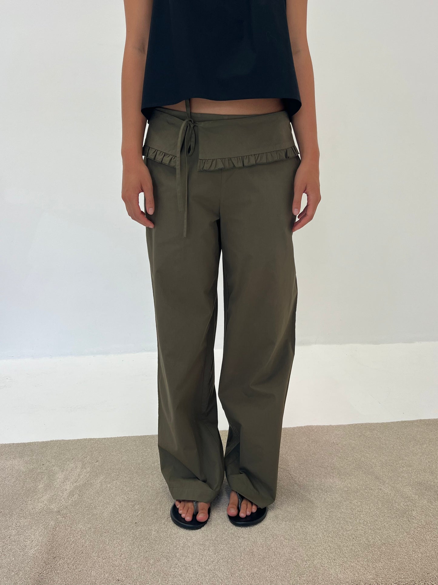 Someone is standing indoors on a light carpet, wearing the Flounce Belt Pant - Khaki featuring a ruffled waistband, paired with a black sleeveless top and black sandals, set against a plain white wall.