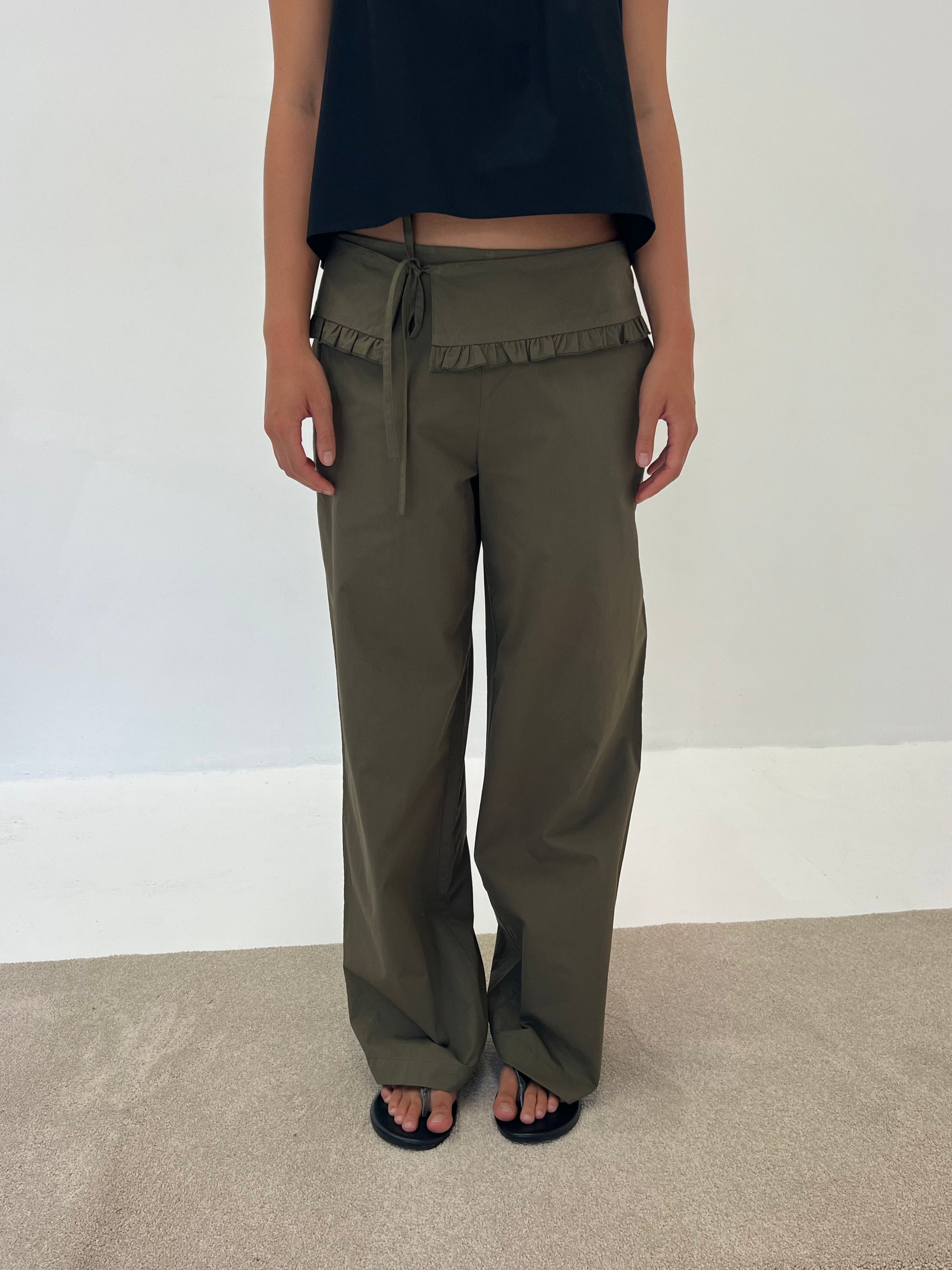 Someone is standing indoors on a light carpet, wearing the Flounce Belt Pant - Khaki featuring a ruffled waistband, paired with a black sleeveless top and black sandals, set against a plain white wall.