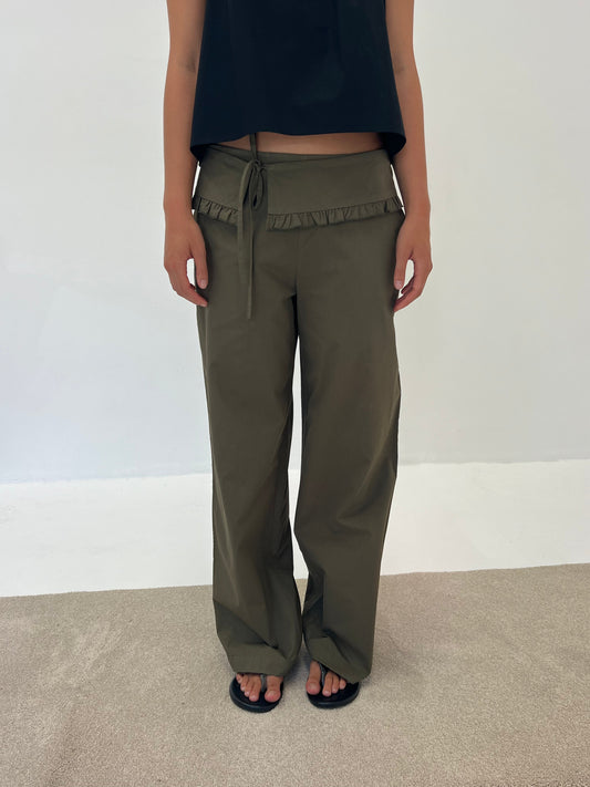 Someone is standing indoors on a light carpet, wearing the Flounce Belt Pant - Khaki featuring a ruffled waistband, paired with a black sleeveless top and black sandals, set against a plain white wall.
