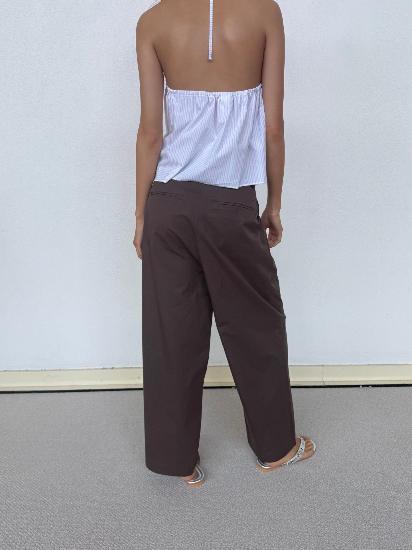A person stands barefoot on a light carpet, wearing the Turn Pant - Cocoa from 0 and a backless, halter-neck white top with thin straps, facing away in front of a plain white wall.