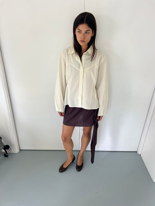 A woman with long dark hair and a neutral expression stands indoors against a white wall, wearing the Cami Overshirt - Citrus Stripe by Deiji Studios, a dark brown asymmetrical skirt with draped detail, and brown flats.