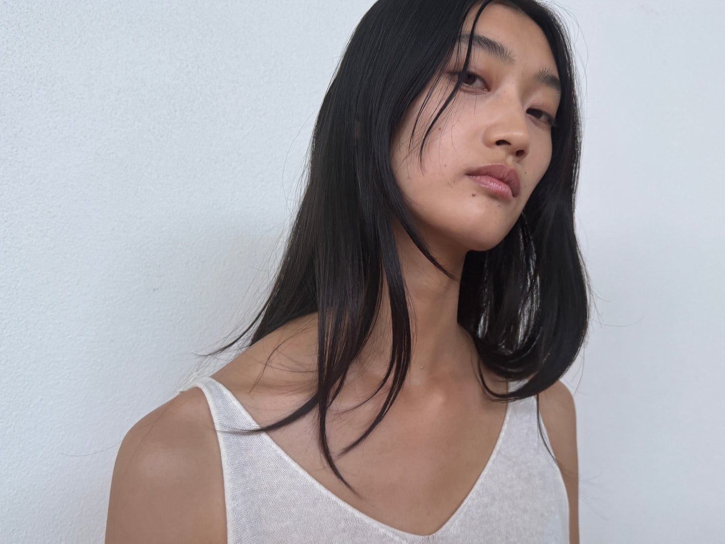 A person with long, straight black hair wears the 0 Rolled Knit Top in Off White and stands against a plain white wall, gazing slightly upwards with a neutral expression.