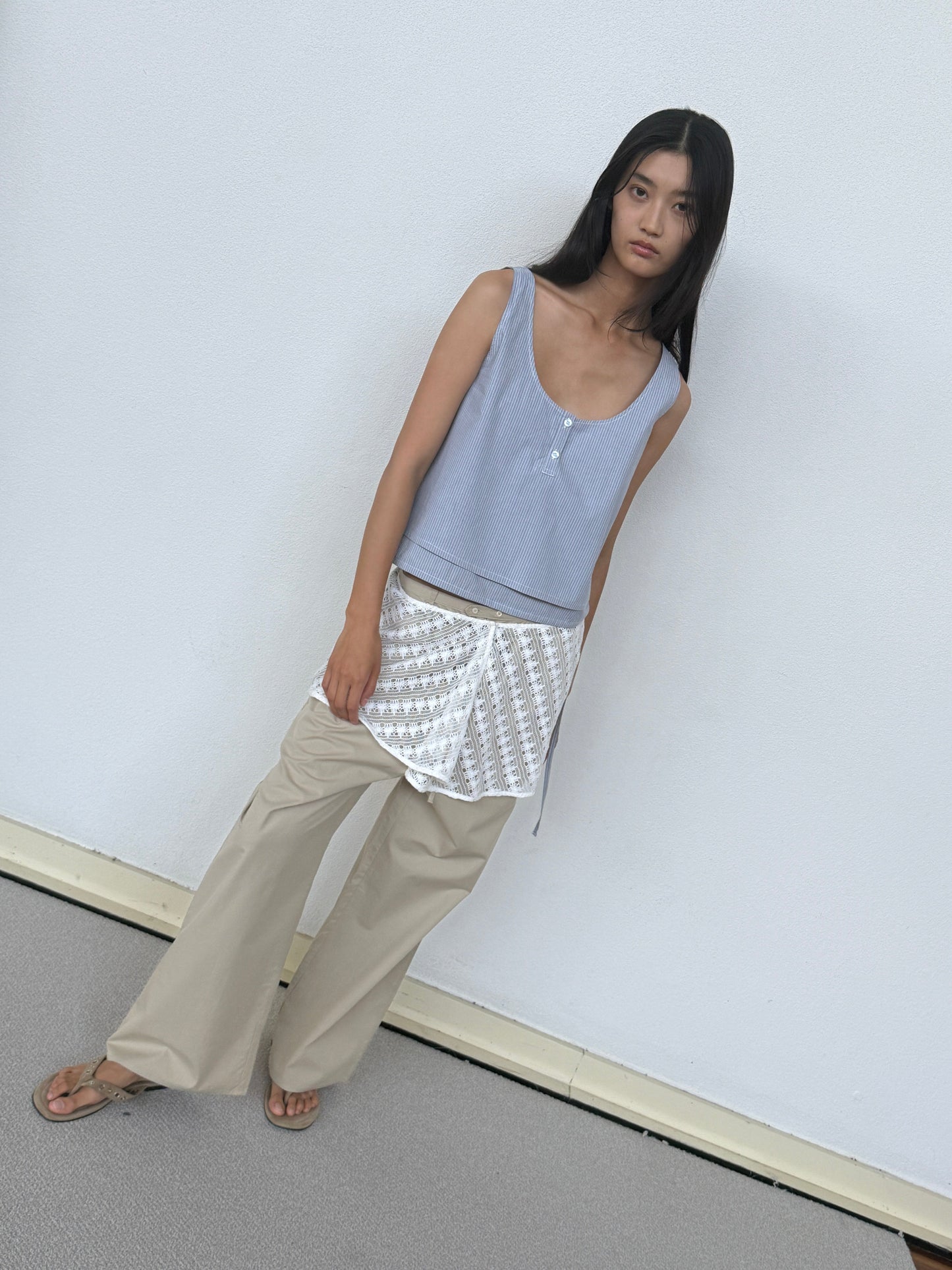 A woman with long dark hair stands against a white wall, wearing the 0 Layered Top - Sky Stripe over a white patterned shirt, beige wide-leg pants, and brown sandals. She looks at the camera with a neutral expression.