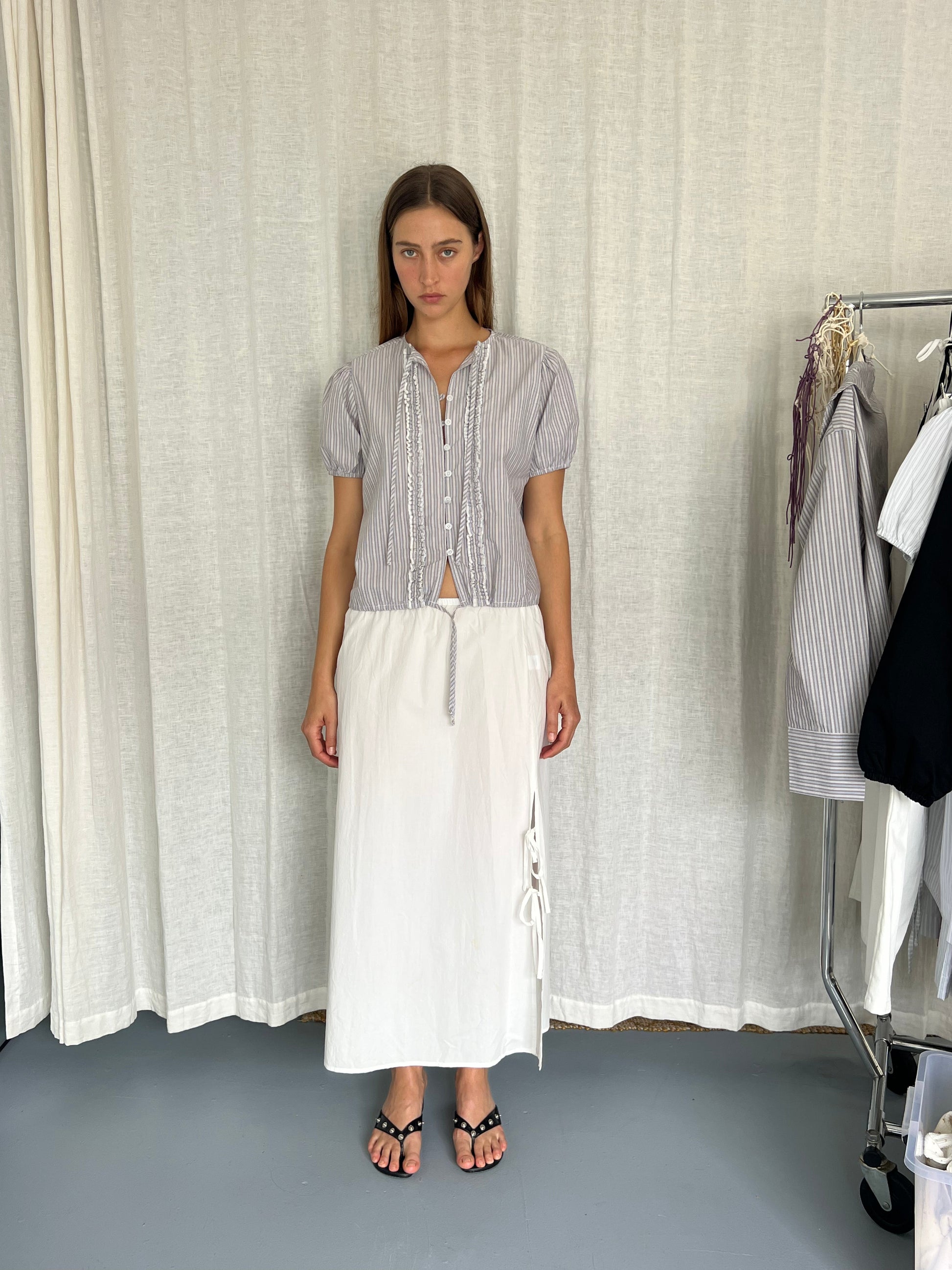 A woman stands indoors wearing the Deiji Studios Double Frill Top in Seas Stripe, paired with a long white skirt with side ties and black sandals. Behind her are hanging shirts and a light curtain backdrop.