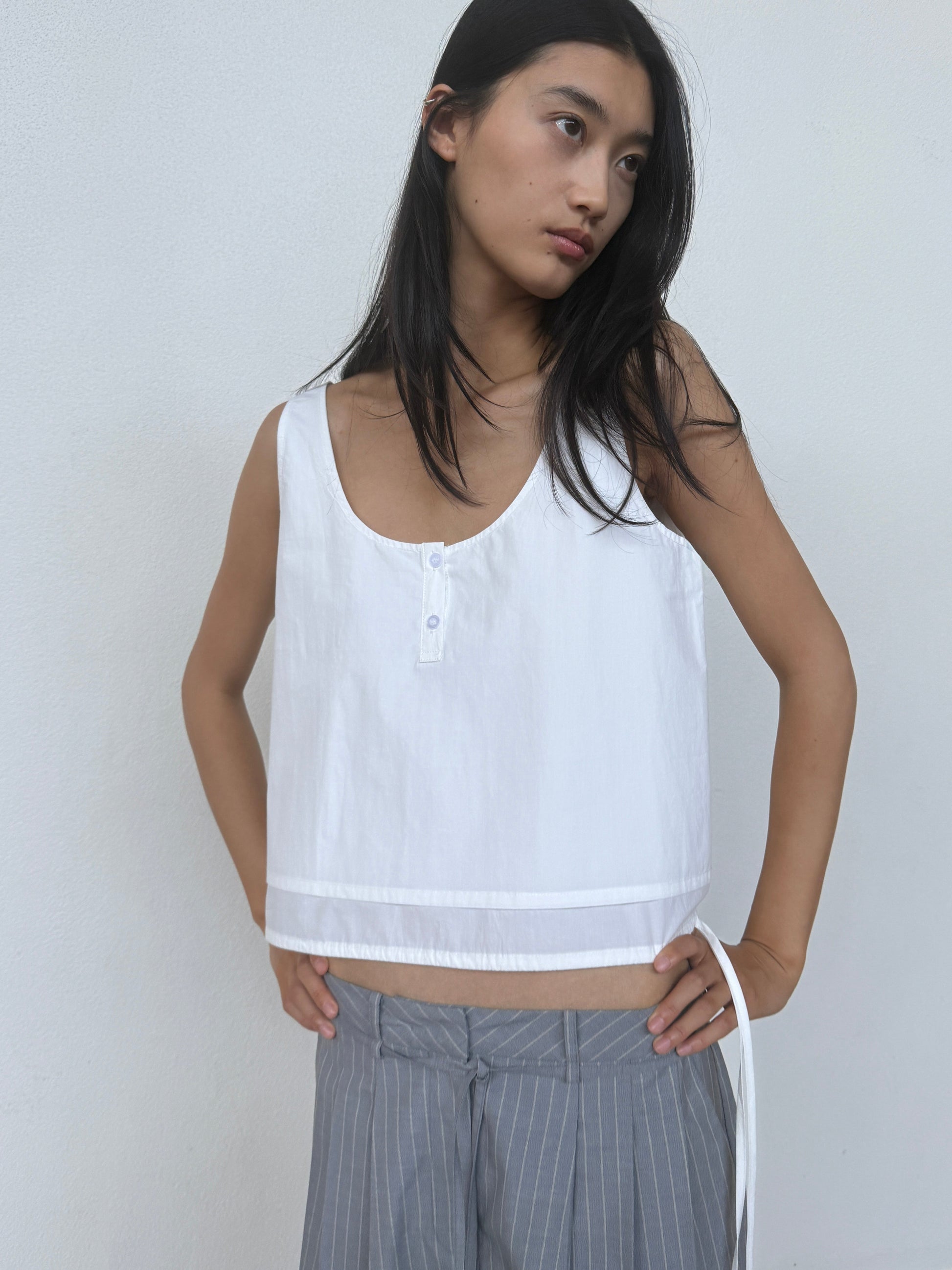 A woman with long black hair wears the Layered Top - White by 0, a sleeveless organic cotton top with buttons, paired with loose gray pinstriped pants, standing against a plain white wall with her hands on her hips.