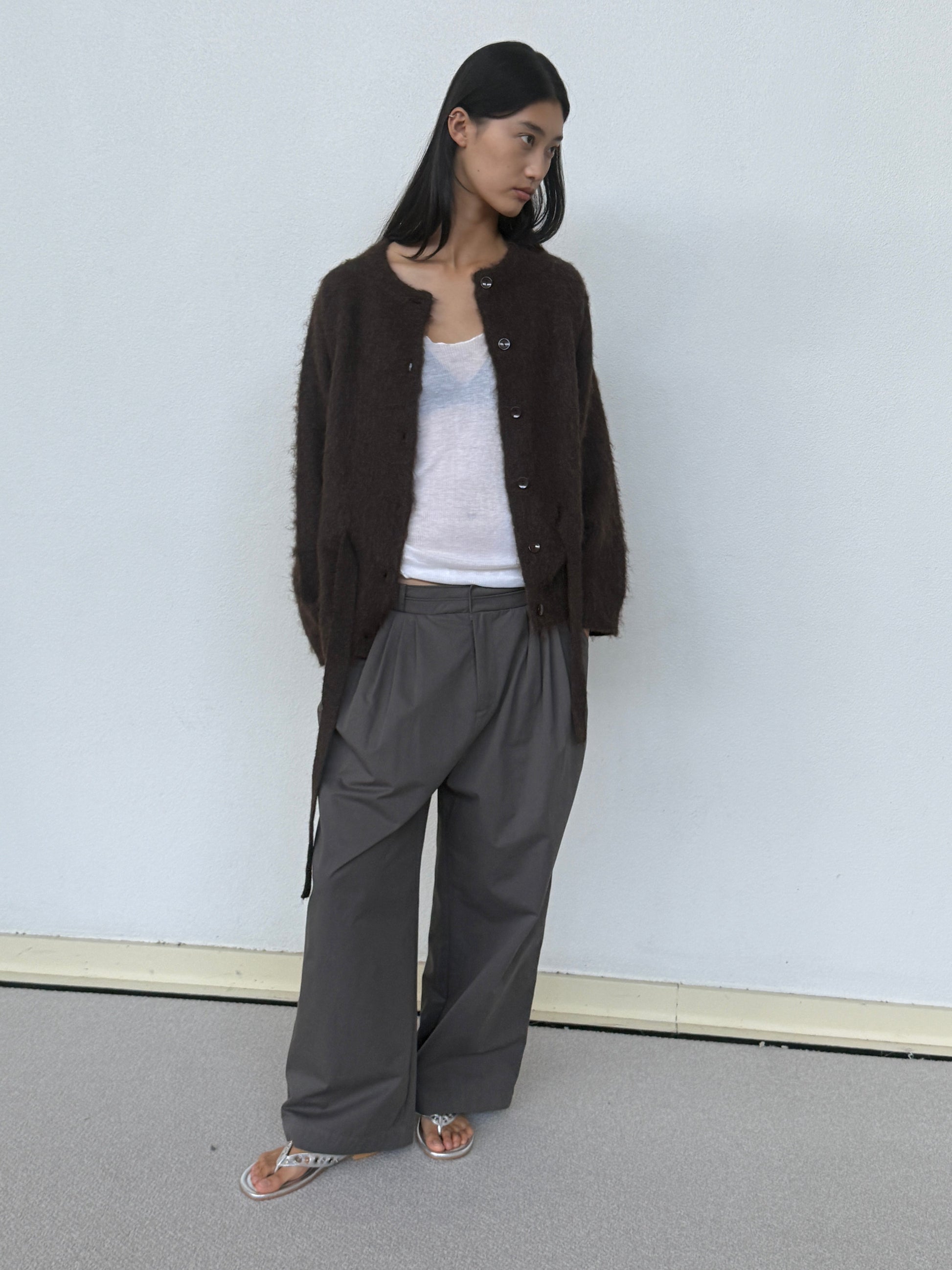 A woman stands against a light wall, wearing a dark brown fuzzy cardigan over a white top and 0’s Two Layer Pant in Charcoal. She completes the look with silver sandals, her long dark hair flowing as she looks left.