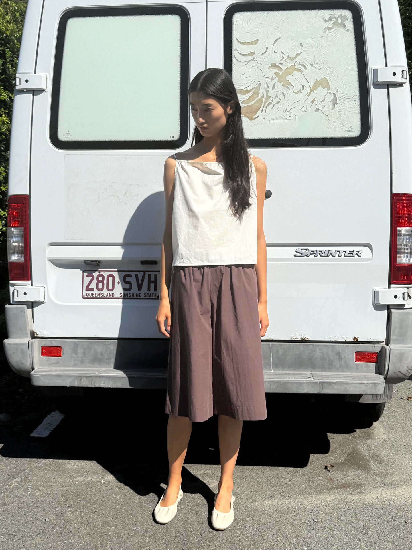 A woman with long dark hair stands in sunlight before a white van, wearing the 0 Long Lounger - Chocolate, an organic cotton relaxed fit sleeveless top and loose brown skirt, finished with beige flats and her head slightly tilted down.