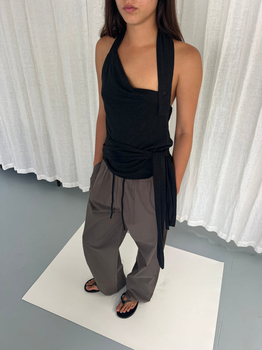 A person stands with hands in pockets, wearing a sleeveless black halter top, Layered Pant - Khaki Check, and black sandals against a light background with white curtains.