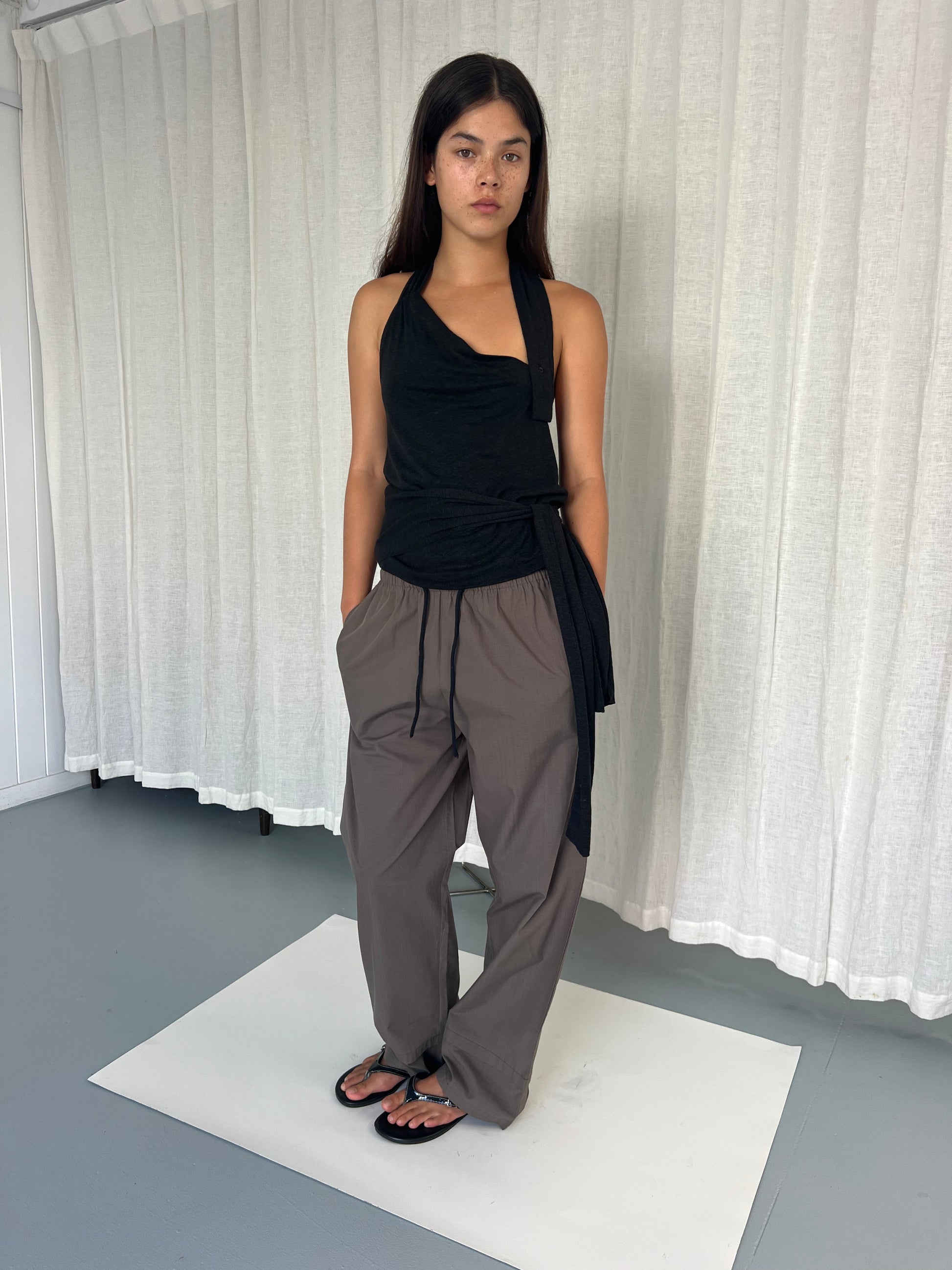 A woman stands on a white mat in front of a white curtain, wearing a sleeveless black asymmetrical top, the Layered Pant - Khaki Check, and black sandals, with her hands in her pockets and a neutral expression.