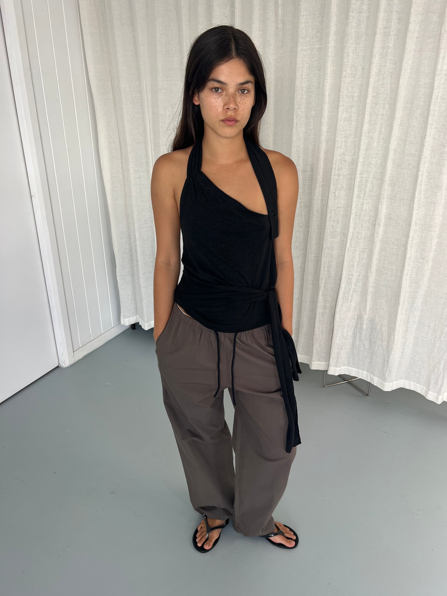 A person stands indoors with hands in pockets, wearing a black halter top, the Layered Pant in Khaki Check, and black sandals. They have long dark hair and a neutral expression, posed before white curtains and a paneled wall.