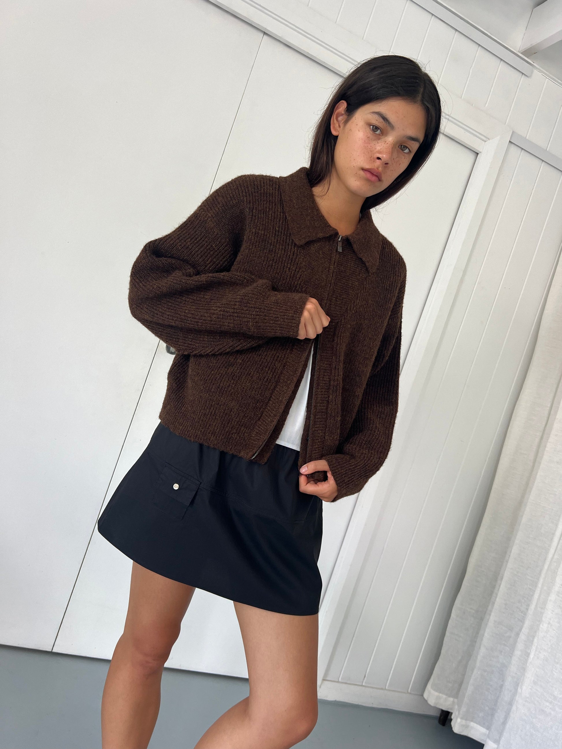 A person with straight dark hair wears the Double Layer Cardigan - Brown from Deiji Studios over a white shirt and black skirt, standing indoors against a white paneled wall and gazing slightly to the side.