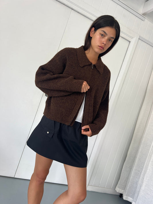 A person with straight dark hair wears the Double Layer Cardigan - Brown from Deiji Studios over a white shirt and black skirt, standing indoors against a white paneled wall and gazing slightly to the side.