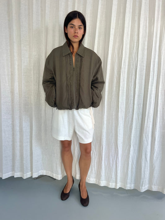 A person stands in front of white curtains, wearing a loose Deiji Studios Pintuck Jacket in Khaki Stripe, white shorts, and dark flat shoes, with hands in their jacket pockets and a neutral expression.