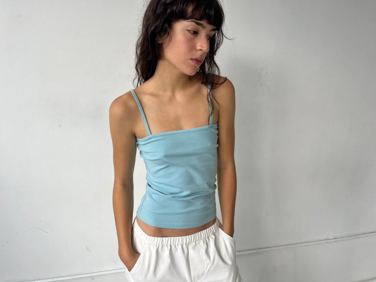 A woman stands against a white wall with hands in her pockets, wearing the Scallop Tank - Sky by Deiji Studios, a fitted ribbed tank with fine straps and scallop trim, styled with the Curve Pant in White.