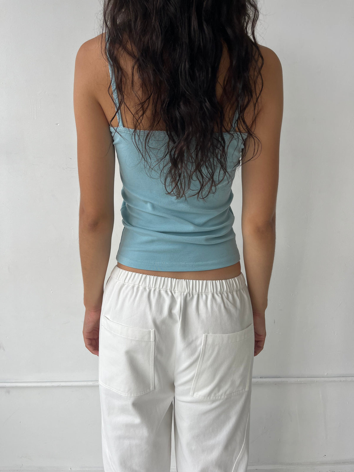 A woman stands facing a white wall, seen from behind, wearing the Scallop Tank - Sky by Deiji Studios, a fitted ribbed tank with fine straps and scallop trim along the straps and back neckline, styled with the Curve Pant in White.