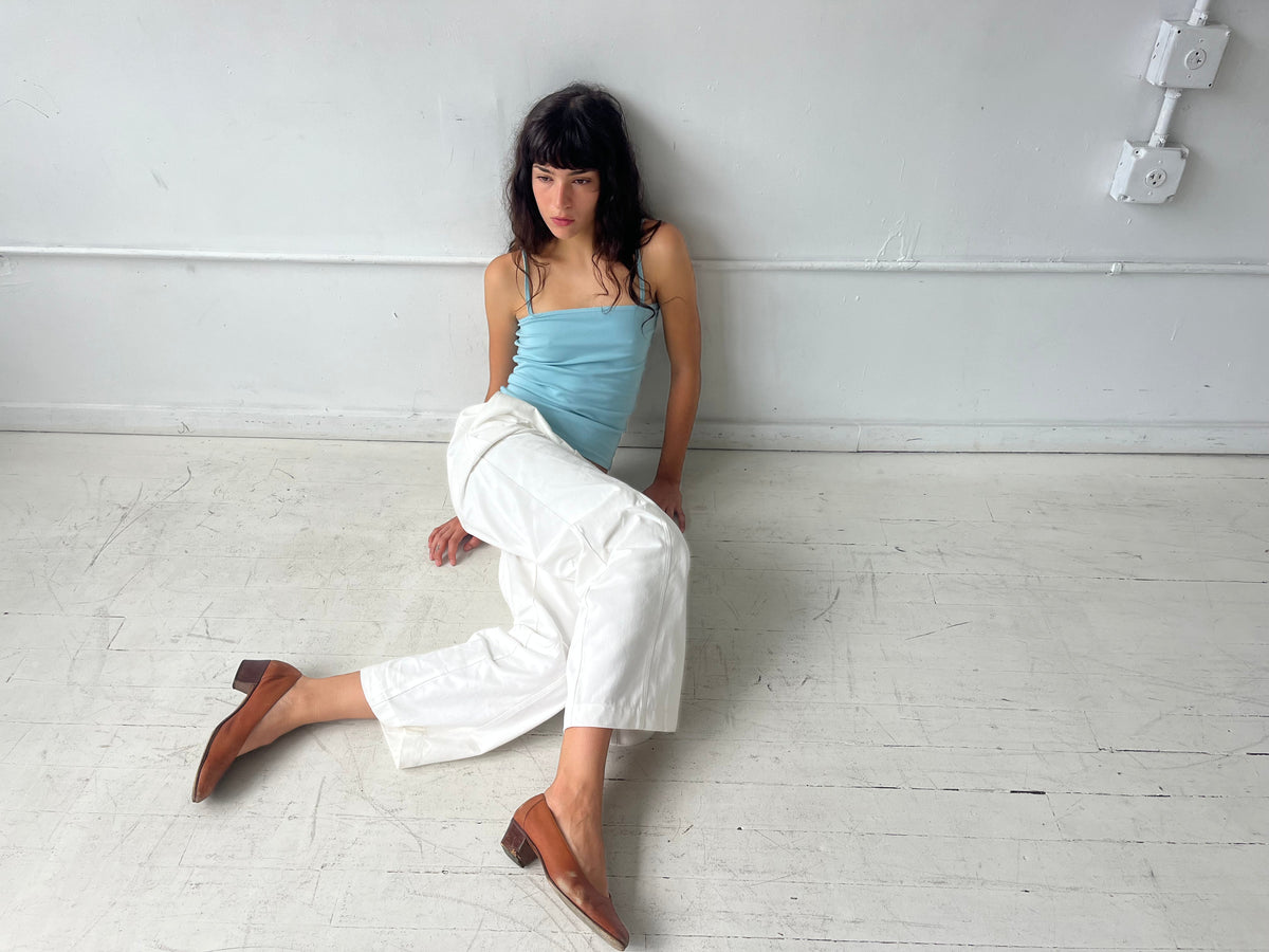 A woman sits on a white studio floor, wearing the Curve Pant - White by Deiji Studios, a softly structured pant with a relaxed fit and curved leg, styled with the Scallop Tank in Sky and brown heels.