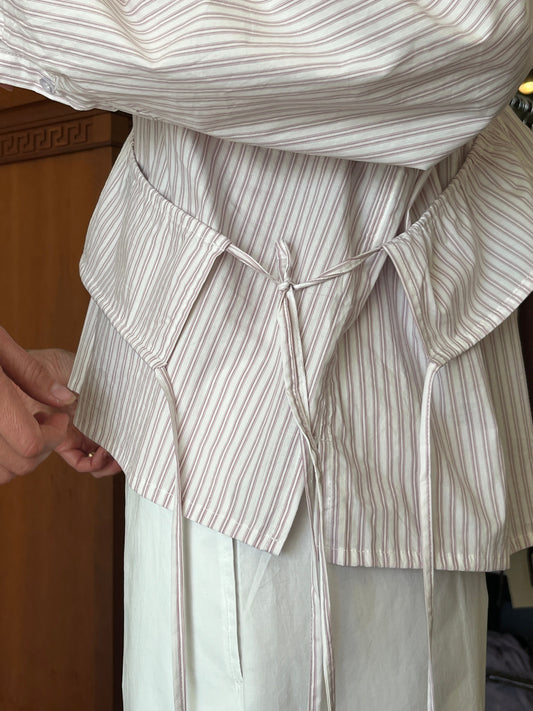 Close-up of a person wearing the Cotton Sash, a light-colored striped shirt with a peplum tie at the waist, paired with white pants. Another hand adjusts the shirt's side near the sash ties.
