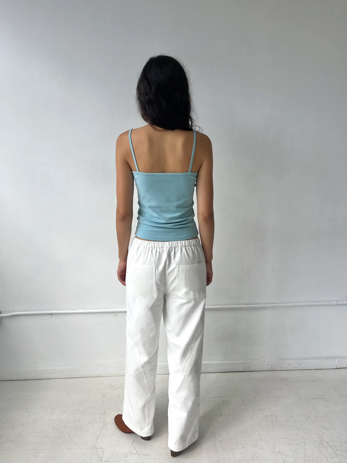A woman stands facing a white wall, seen from behind, wearing the Curve Pant - White by Deiji Studios, a softly structured pant with a relaxed fit and back patch pockets, styled with the Scallop Tank in Sky.